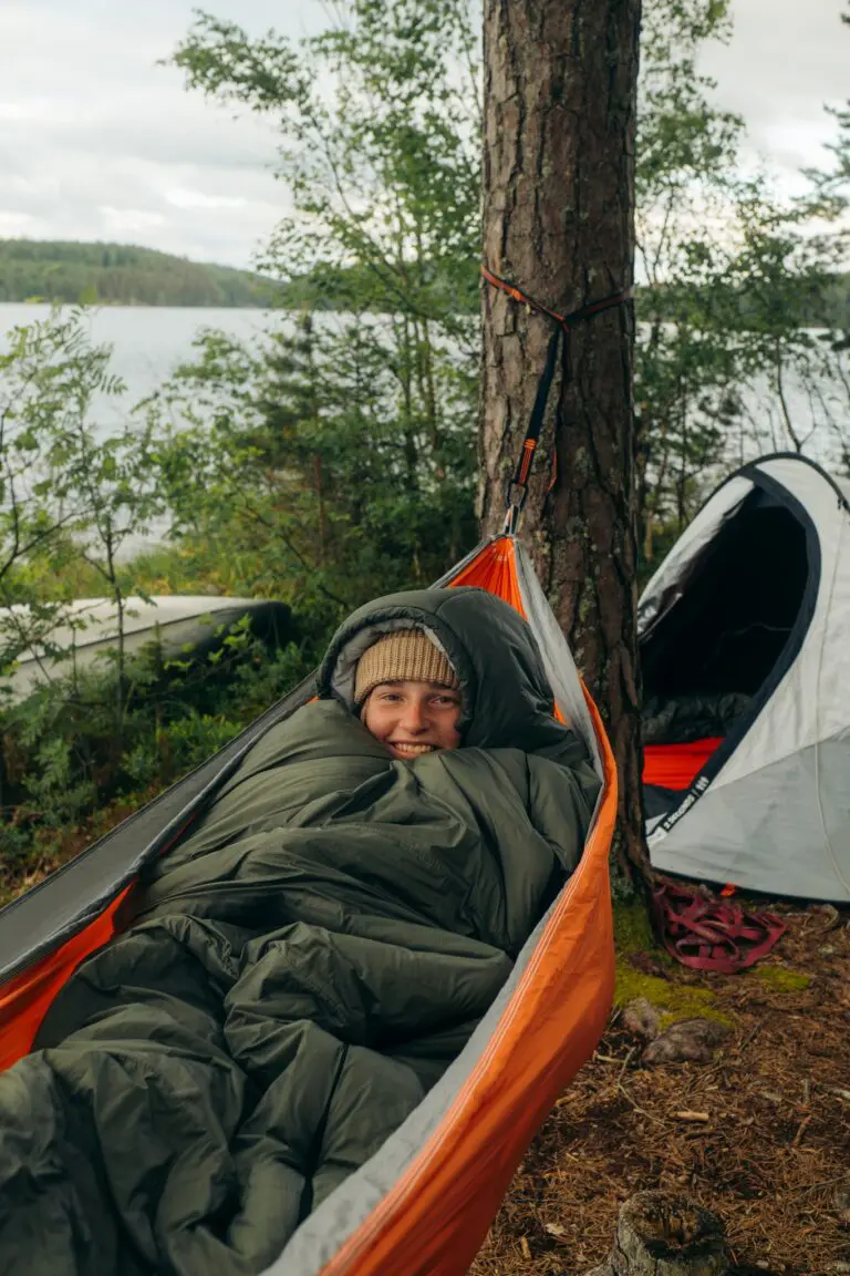 person smiling in hammock by lake with tent